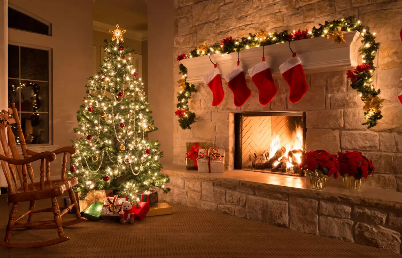 Sapin de Noël illuminé et décoré avec des guirlandes dorées et des ornements rouges, entouré de cadeaux, près d'une cheminée ornée de chaussettes de Noël rouges et d'une guirlande lumineuse, créant une ambiance chaleureuse et festive.